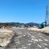 A vast construction site with heavy machinery and building materials scattered throughout. An unfinished road stretches through the center. In the background, mountains and a residential area can be seen under a clear blue sky.