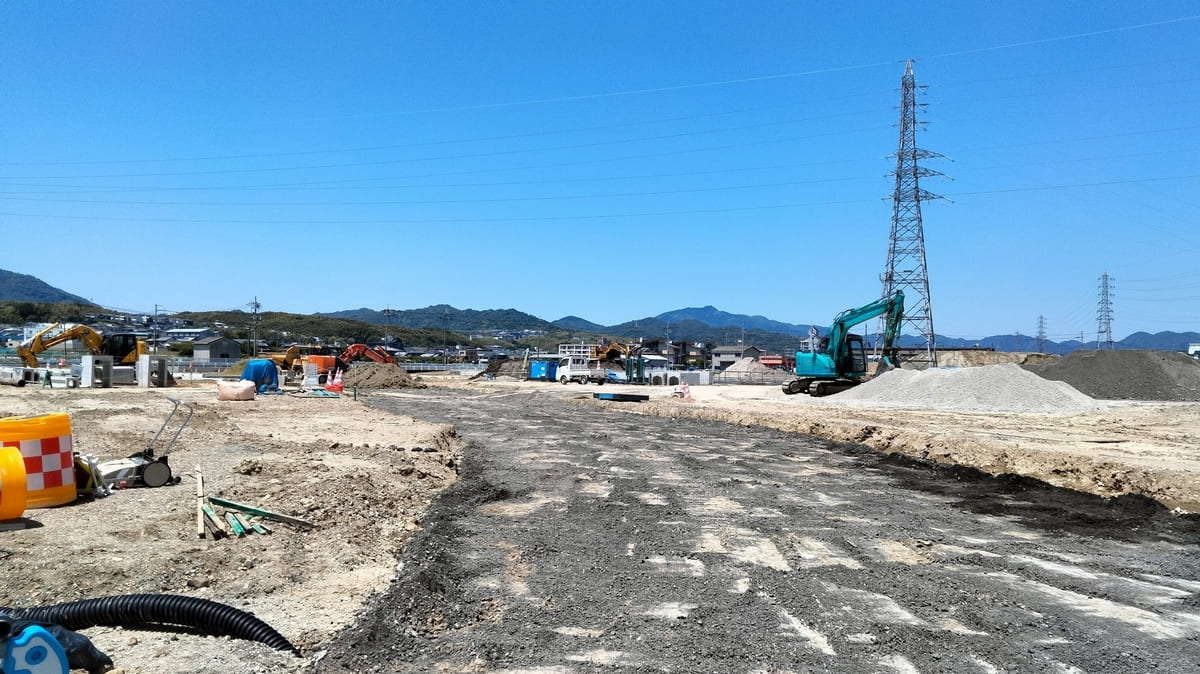 A vast construction site with heavy machinery and building materials scattered throughout. An unfinished road stretches through the center. In the background, mountains and a residential area can be seen under a clear blue sky.
