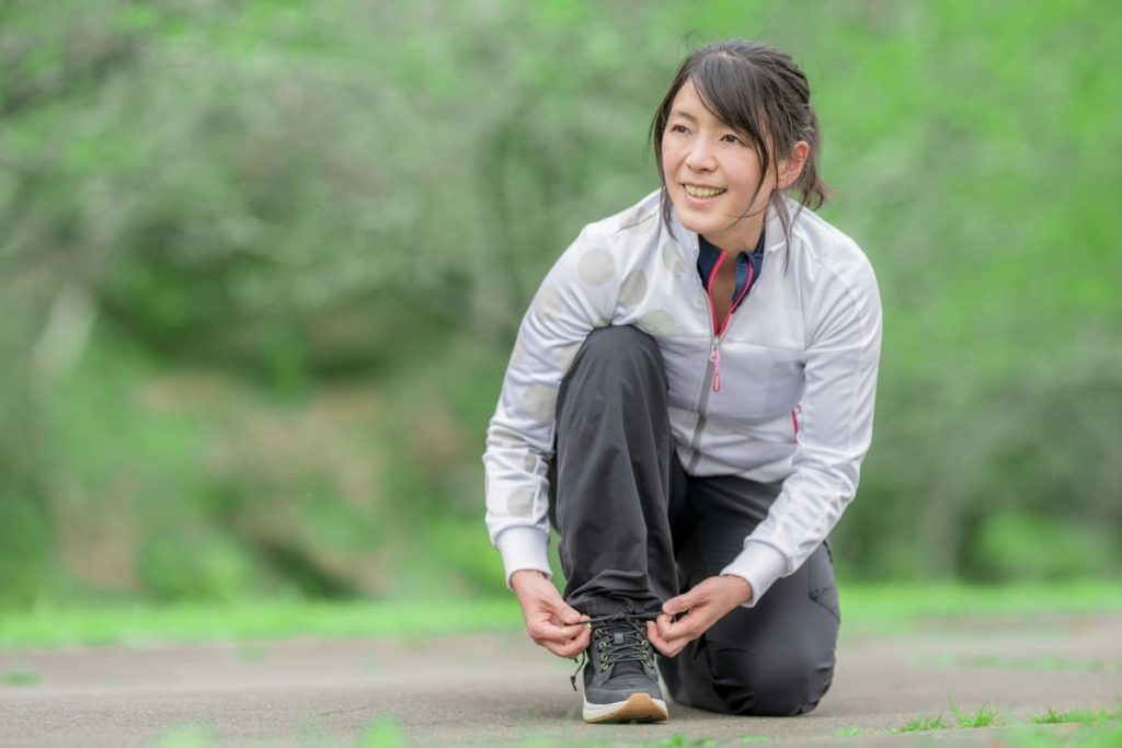 屋外の緑豊かな環境で、笑顔の女性がしゃがみ込みながら靴ひもを結んでいる。スポーツウェアを着用し、これから運動を始める様子がうかがえる。リラックスした雰囲気が漂う。