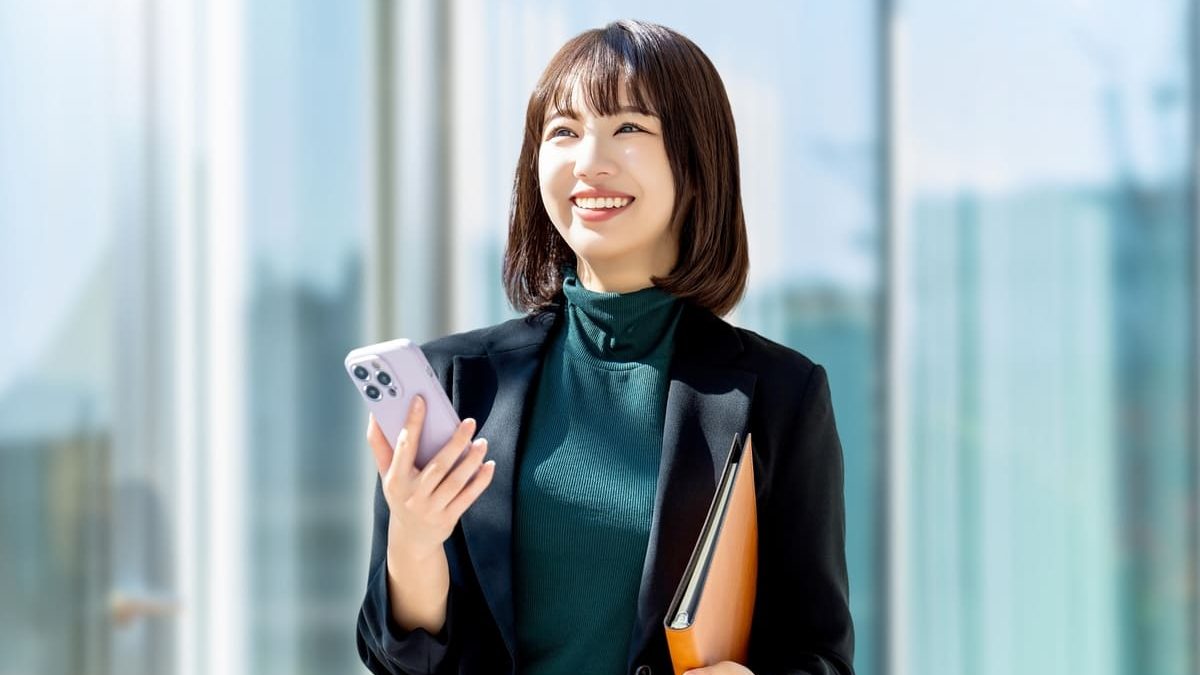 黒いブレザーと緑のタートルネックを着た若いビジネスウーマンが、近代的なガラス張りの建物の前で微笑みながらスマートフォンとオレンジ色のフォルダーを持っている。