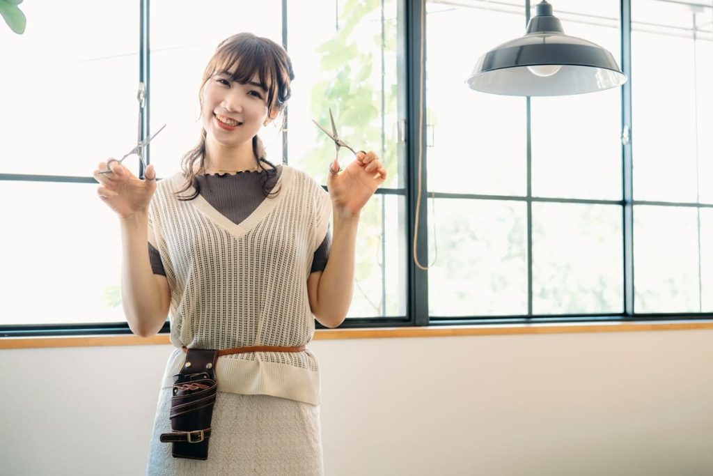 笑顔の美容師が、両手にハサミを持ち、明るくモダンなサロンの中でポーズを取っている。ベージュのベストをダークカラーのトップスの上に着用し、腰にはレザーのツールベルトをつけている。