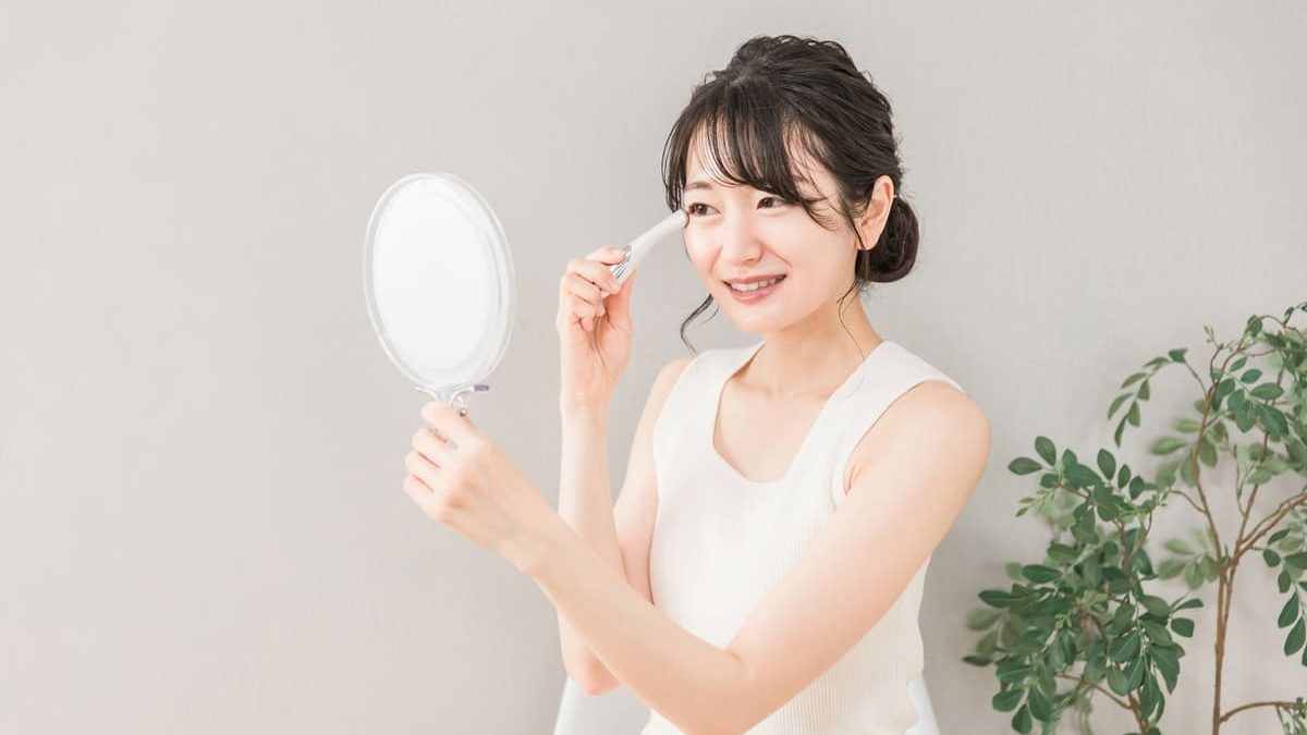 A smiling woman applying a beauty stick to her eye area while holding a hand mirror. She is wearing a white sleeveless top, and a potted plant is visible in the background.