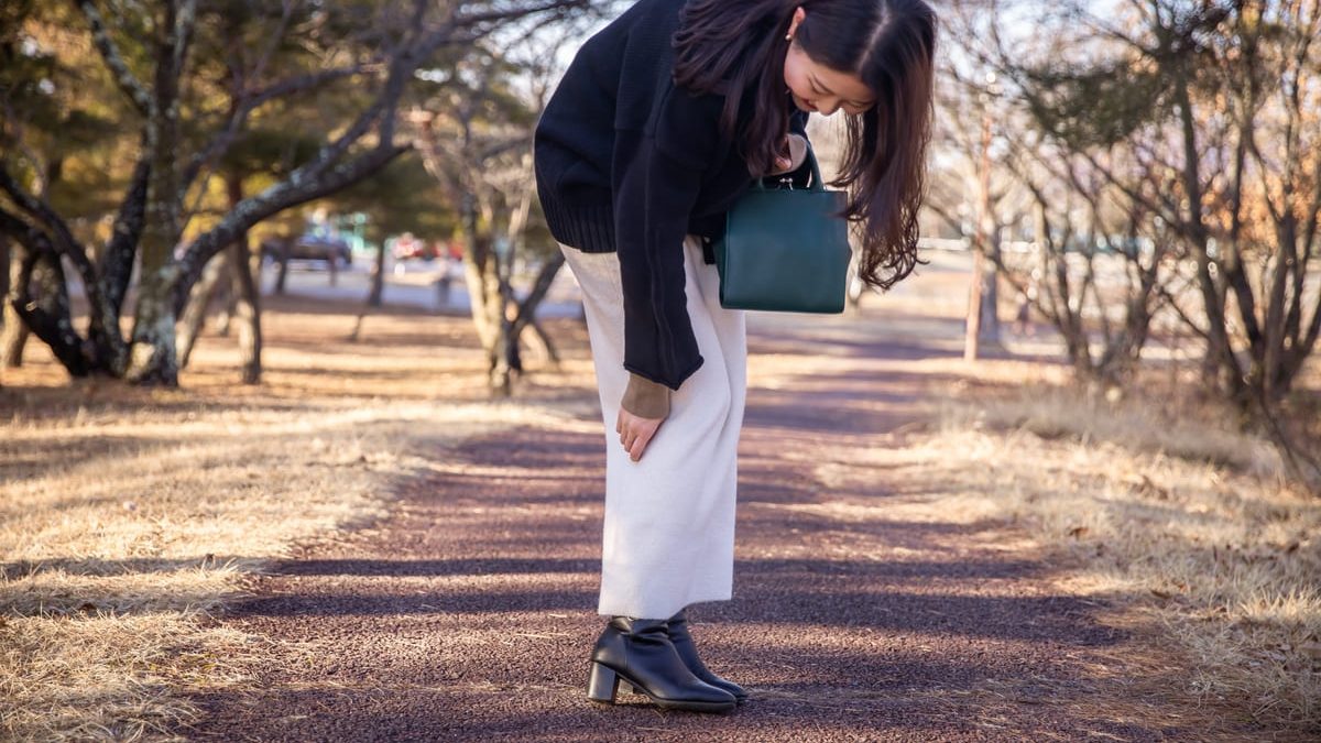 A woman in a black sweater and white pants bends down to touch her knee while walking on a park path. She holds a dark green handbag, and the surroundings have trees and dried grass.