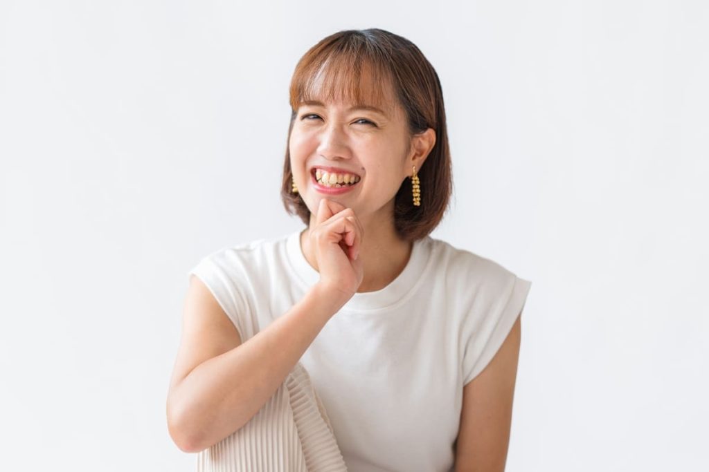 白いノースリーブトップスとゴールドのイヤリングをつけた女性が、顎に手を添えて明るく微笑んでいる。背景はシンプルな白色。