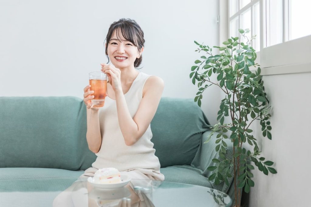 ファに座り、笑顔でアイスティーを持つ女性。ガラスのテーブルにはケーキが置かれており、背景には観葉植物と窓が見える。