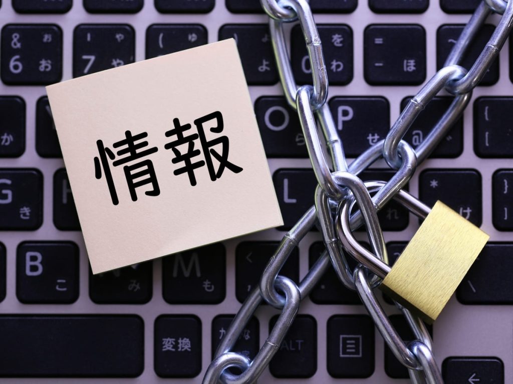 「情報」と書かれた紙がパソコンのキーボードの上に置かれ、鎖と南京錠でロックされている。情報セキュリティやデータ保護の概念を表現したイメージ。
