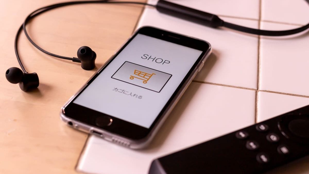A smartphone screen displaying "SHOP" with a shopping cart icon, surrounded by earphones and a remote control, representing online shopping.