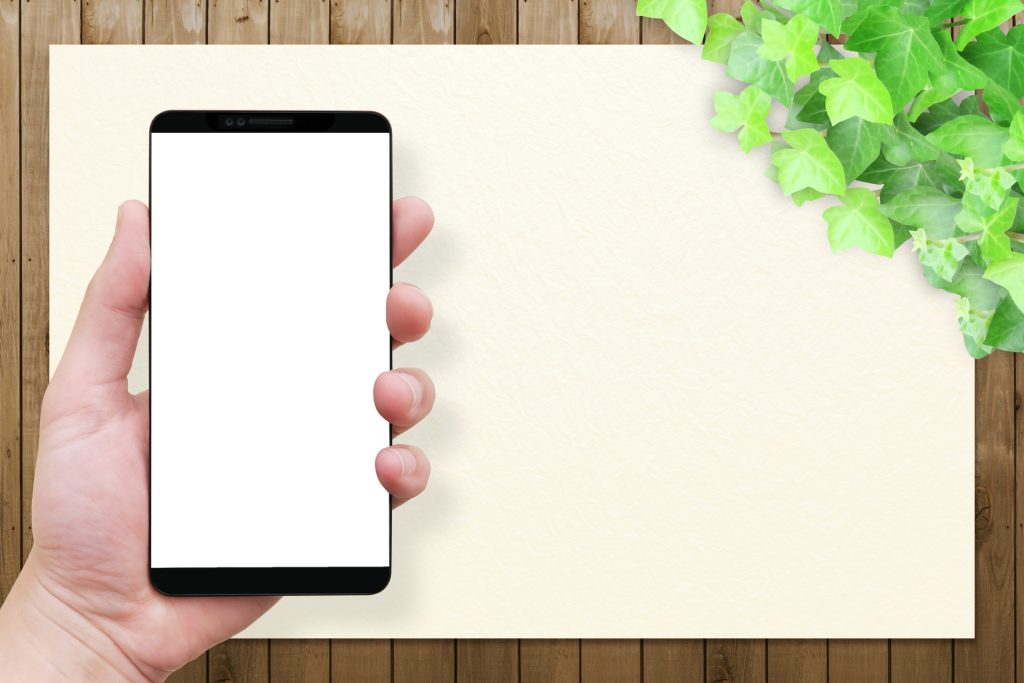 手に持ったスマートフォンと白い紙、観葉植物が飾られた木目調の背景の画像。