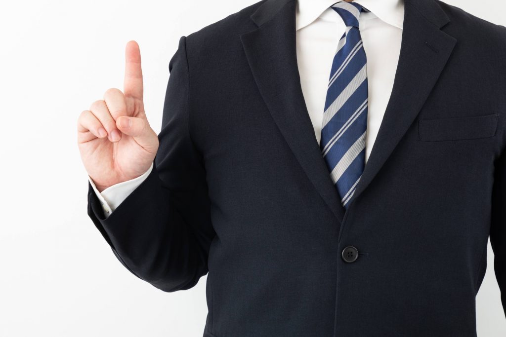 紺色のスーツとストライプのネクタイを着用した男性が、白い背景の前で人差し指を立てている様子。