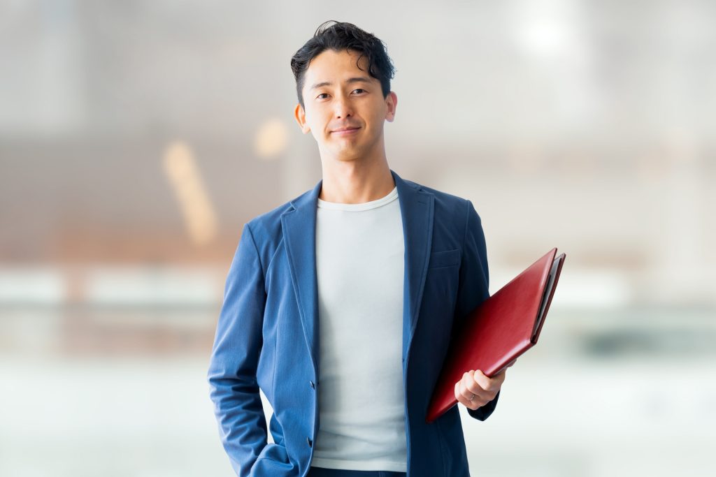 ビジネスカジュアルな青いジャケットと白いTシャツを着用し、赤いファイルを持つ男性がオフィスの背景の前に立っている様子。
