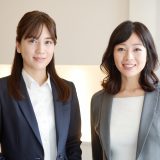 Two women wearing business suits are standing side by side in an office. The image represents a mental coach and a woman receiving coaching.