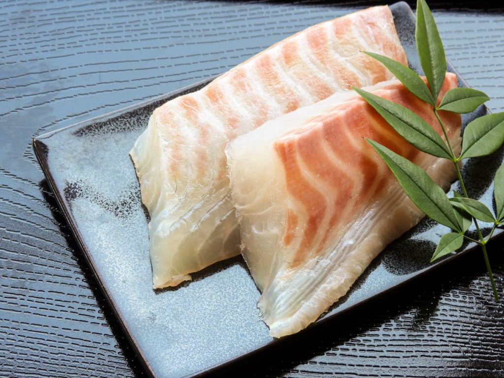 黒い皿に盛り付けられた真鯛の切り身と葉の写真。薄く切って鯛めしの材料にする。