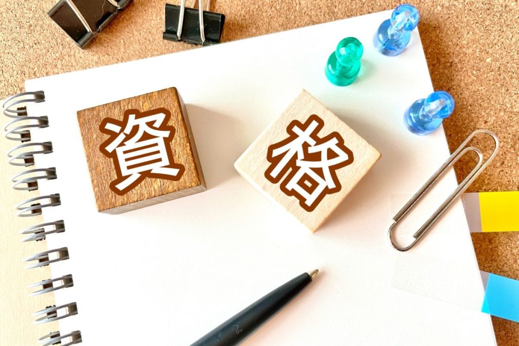 資格の文字が書かれた木製ブロックとノート、文房具が並ぶ机の上