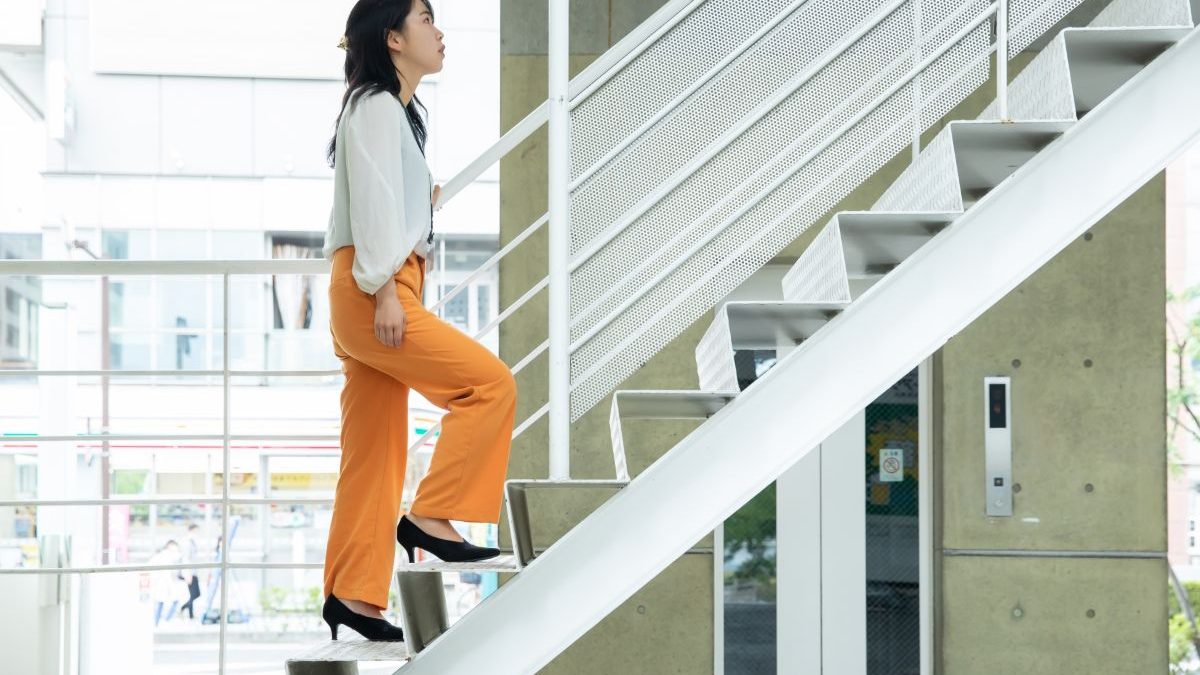 A woman in a white blouse and orange pants is walking up the stairs indoors with a focused expression. The scene evokes themes of health, fitness, and a positive lifestyle.