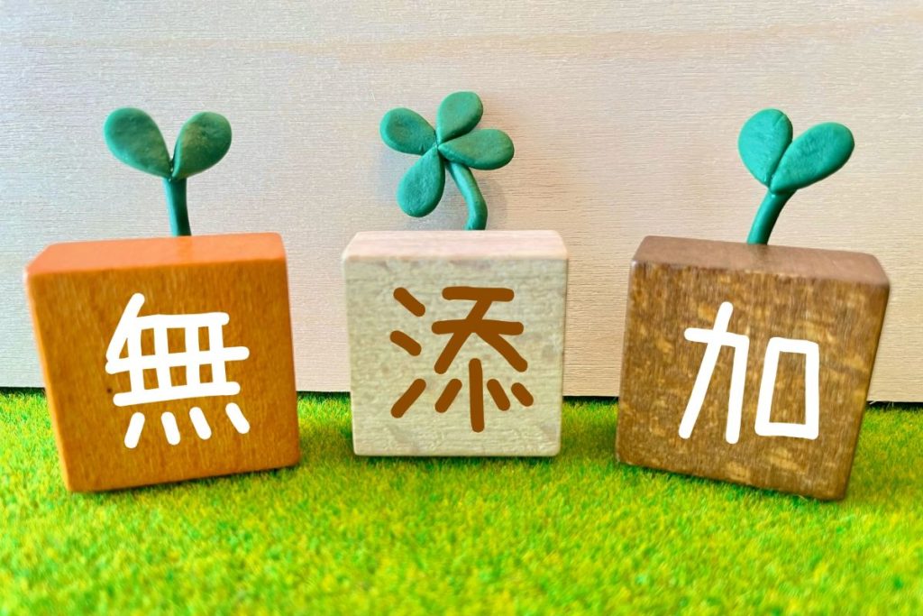 「『無添加』という文字が書かれた3つの木のブロックにクローバーの芽が生えたナチュラルなデザイン。安心・安全なペットフードや自然志向の商品を連想させるイメージ。