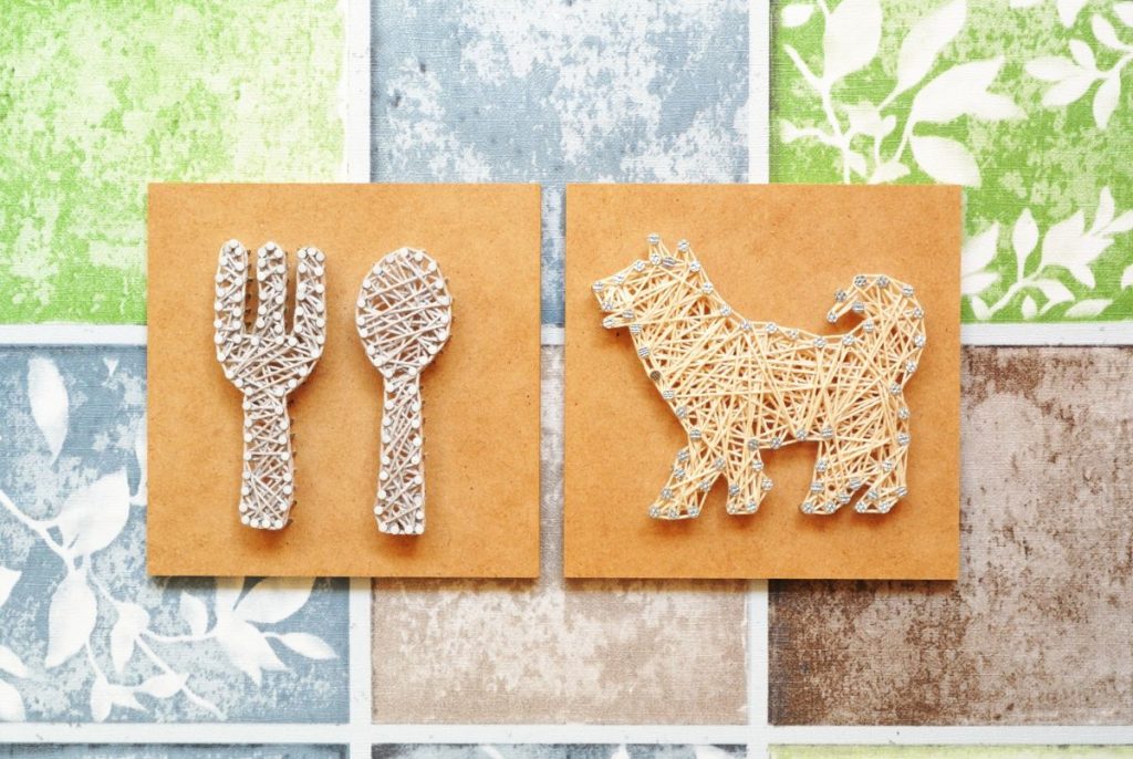 カラフルな背景の上に配置された、糸で作られたスプーン・フォークと犬の形のアート作品。ペットと食事の関係や、手作りご飯、愛情を込めたケアを象徴するクラフト風のイメージ。