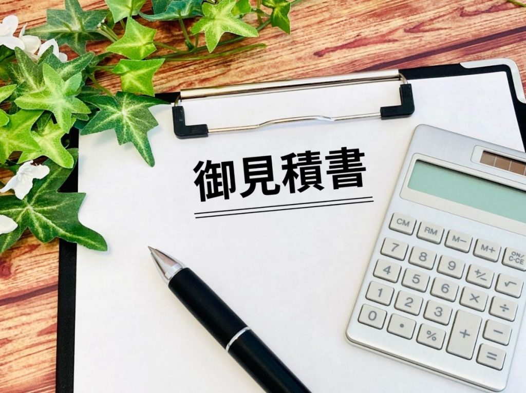 クリップボードに「御見積書」と書かれた用紙が乗っており、隣に電卓と黒いボールペン、さらに左上には観葉植物の葉が添えられている。木目調のテーブルの上で、見積書作成や費用計算のイメージを表現した写真。