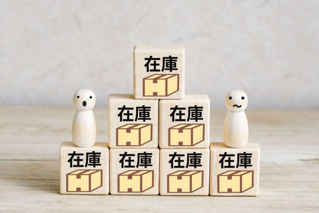 在庫と書かれたブロックを積み上げている様子と、驚いた表情の人形。過剰在庫や在庫管理のイメージ。