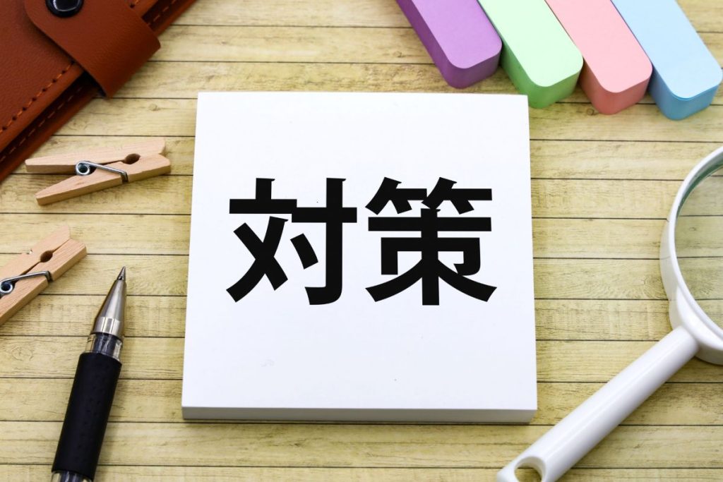 木目のデスクの上に「対策」と大きく書かれた白いメモが置かれている。周囲にはカラフルな付箋やクリップ、ペン、虫眼鏡などが並び、問題解決やビジネス戦略の策定をイメージさせる構成。