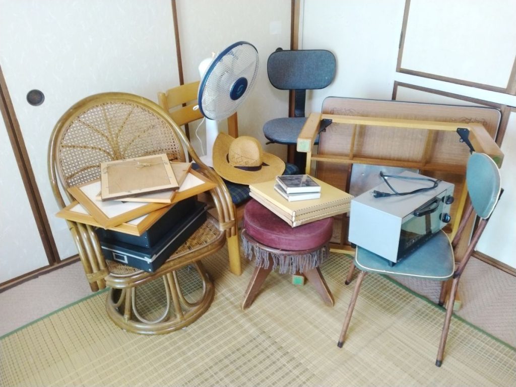畳の部屋に、椅子や扇風機、額縁、帽子、本、家電などの不要品がまとめて置かれている。引っ越しや片付け、不要品回収のイメージを伝える写真。