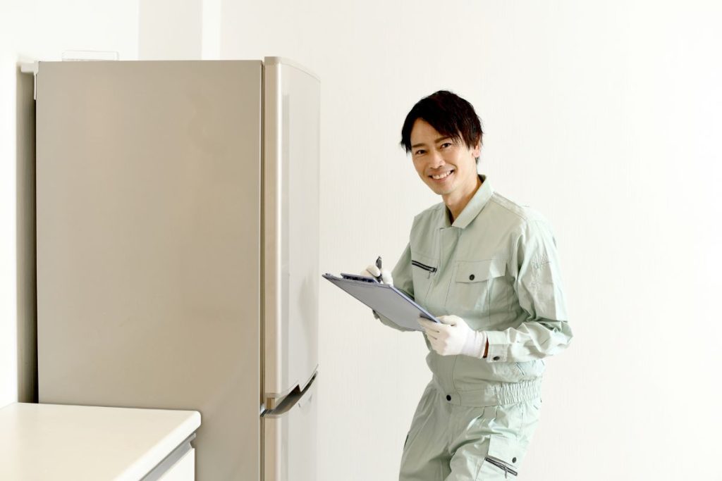 作業服を着た作業員が、冷蔵庫の前でクリップボードを持ってメモを取っている。家電の回収や見積もり、引っ越し作業のイメージを伝える写真。

