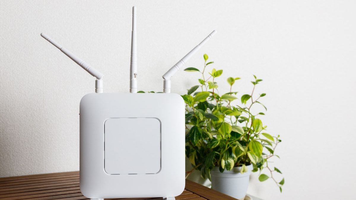 A white Wi-Fi router with three antennas is placed on a wooden table. A potted plant in the background adds a clean and natural atmosphere to the interior setting.