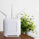 A white Wi-Fi router with three antennas is placed on a wooden table. A potted plant in the background adds a clean and natural atmosphere to the interior setting.