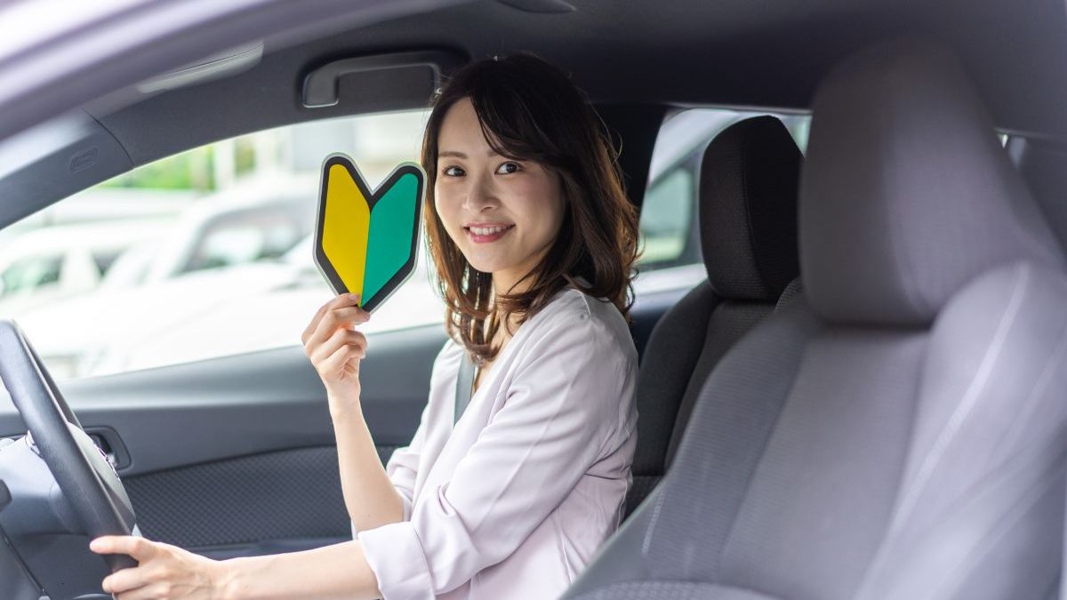 A woman sitting in the driver’s seat of a car holds a green and yellow beginner driver’s mark (shoshinsha mark) and shows it through the window. The background shows a parking lot, indicating that she is a new or inexperienced driver.