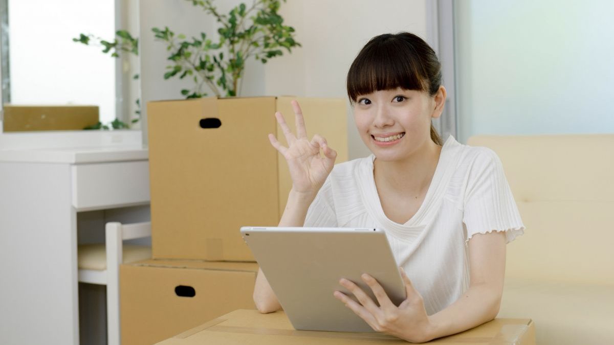 A smiling woman making an OK gesture while using a tablet in front of cardboard boxes. Represents inventory management.