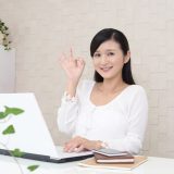 A smiling woman working on a computer at her desk is making an "OK" sign with her left hand, giving the impression that her online business is going well.