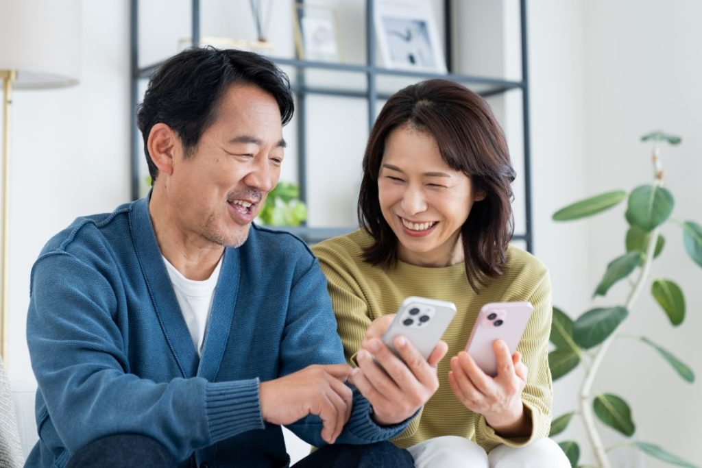 スマートフォンを手に取り、笑顔で会話する中年の日本人夫婦の画像。テクノロジーを通じた日常の楽しみや情報共有を表現。