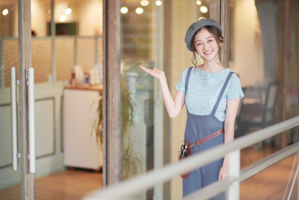 女性の美容師が笑顔で美容室の入り口で客を案内する様子。