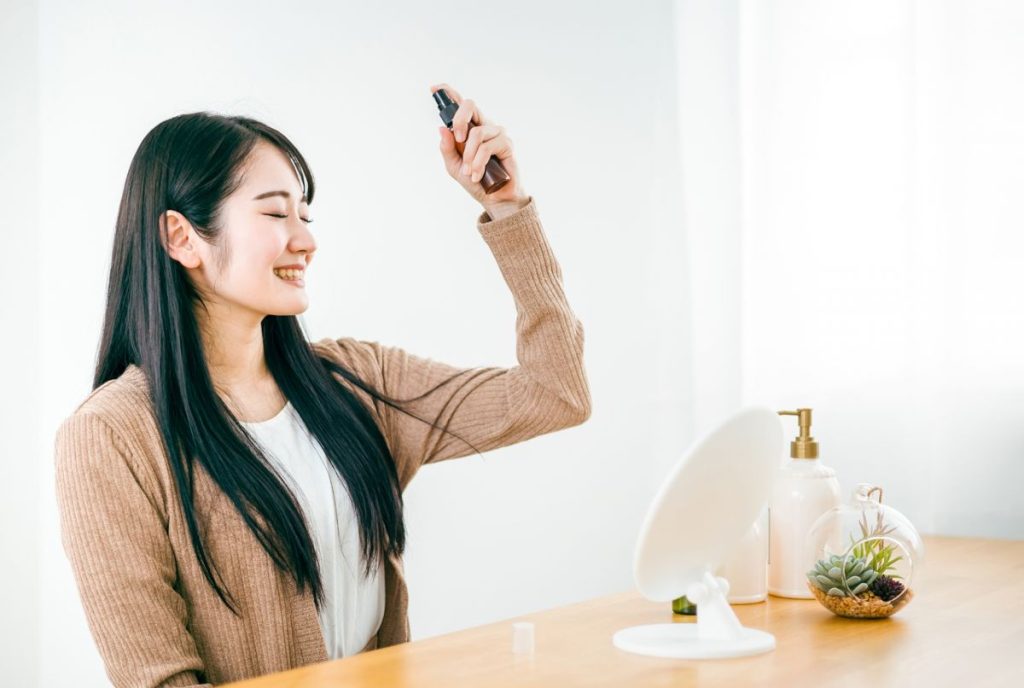 鏡の前でヘアミストを髪にスプレーしている笑顔の女性。静電気対策やヘアケアの一環として使っている場面。