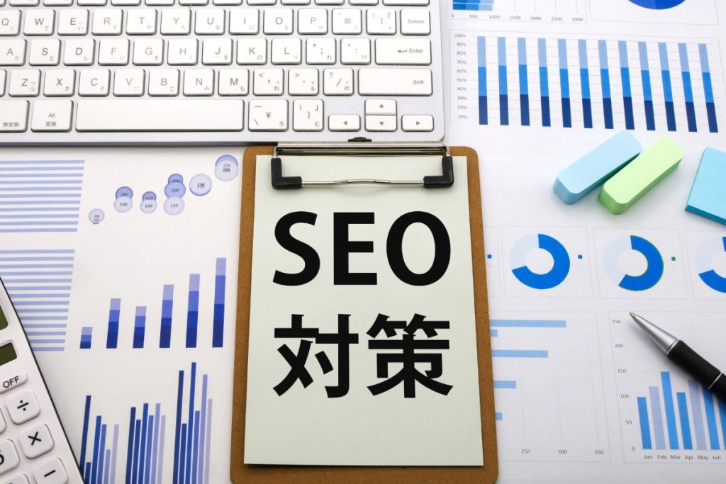 「SEO対策」と書かれた紙の周りにパソコンのキーボードや電卓、ペン、附箋が置かれている。背景一面に売上データが表示されている。