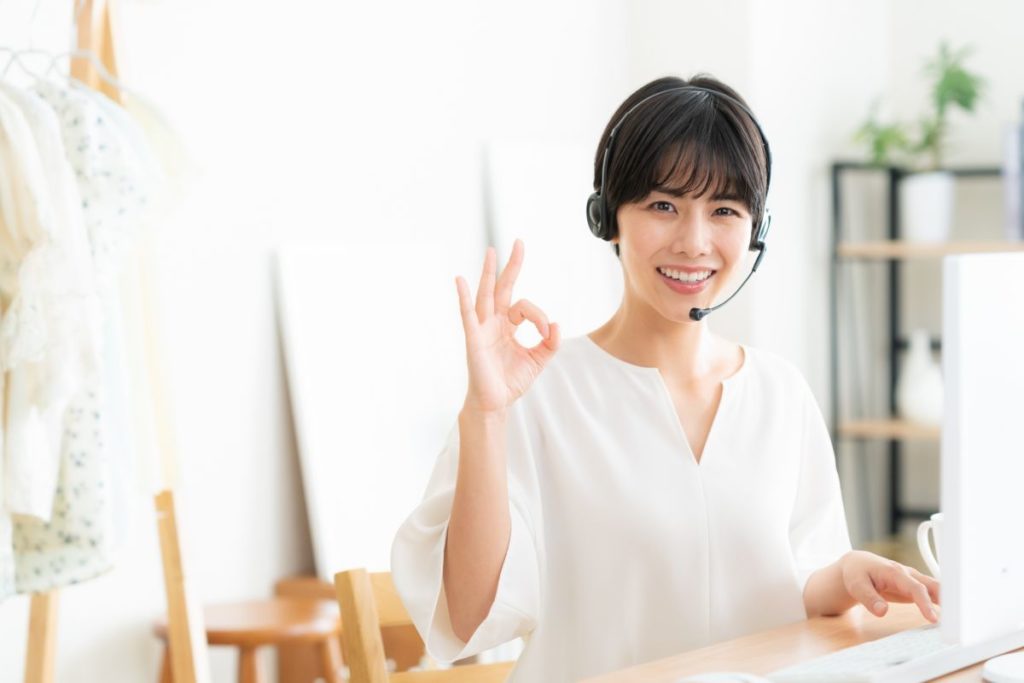 清潔感のある身だしなみの女性が、リビングでヘッドフォンを付けてオンライン会議に出席している様子。笑顔でオッケーサインをしている。