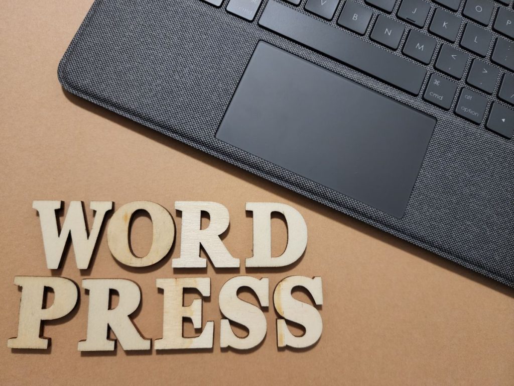デスクにノートパソコンが置かれており、そばには「WORD PRESS」のアルファベットピースが並べられている。アフィリエイトサイトの制作準備をしているイメージ。