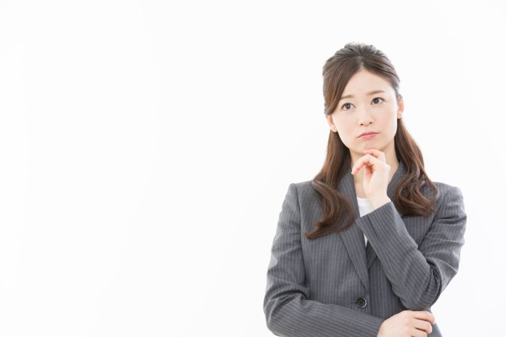 スーツ姿の女性が腕を組んで考え込む様子。コーチングやカウンセリングの選択に迷っている場面を表現。