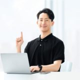 A confident male coach sitting in front of a laptop, raising his finger as he offers guidance. Represents trust and insight in the coaching process.