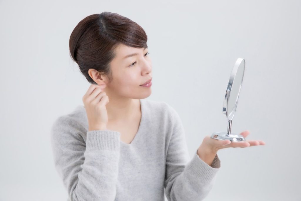 鏡を見ながら耳のツボを確認している女性。耳ツボジュエリーを自分で貼るときの実践シーンを表現した画像。