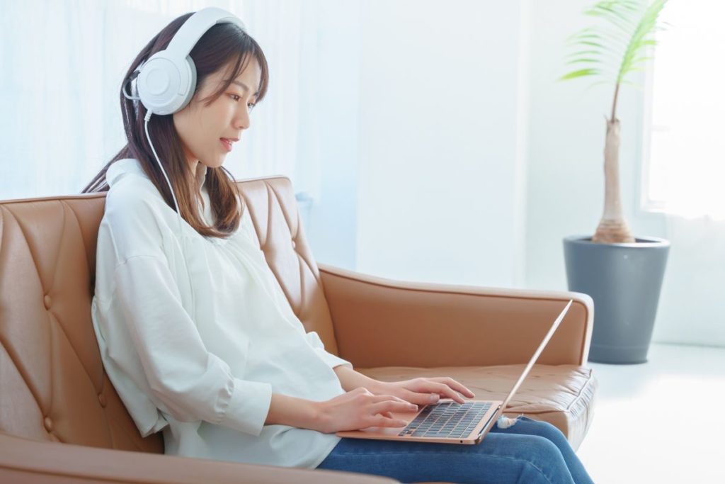 ソファに座った女性がヘッドフォンをつけ、オンライン学習でスキルアップに取り組んでいるイメージ。