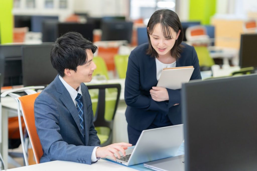 オフィスで新人社員が先輩社員にパソコンを見ながらわからないことを質問しているイメージ。