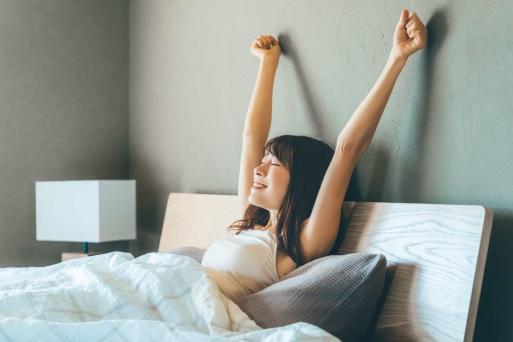 ベッドの上で気持ちよさそうに伸びをし、良く眠れた様子の女性の画像。