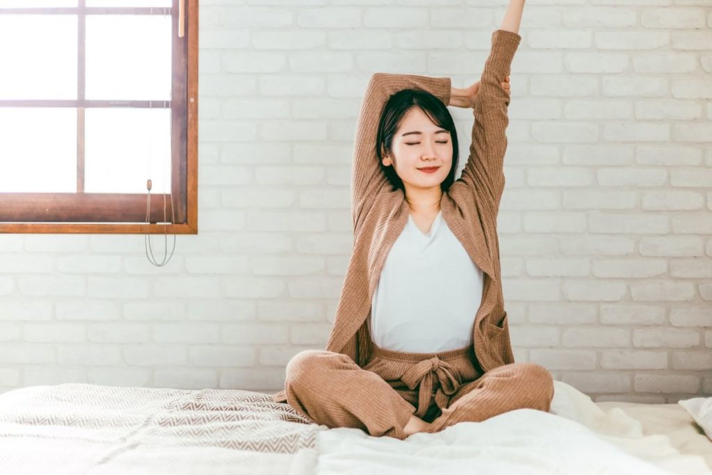 朝の光の中でベッドの上で気持ちよさそうに深呼吸しながら伸びをしている女性のイメージ。