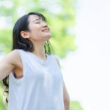 An image of a woman outdoors surrounded by greenery, stretching out her arms and taking a refreshing deep breath.