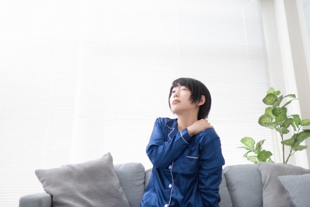 パジャマ姿で肩に手を当て、体調の悪さに悩んでいる様子の女性のイメージ。