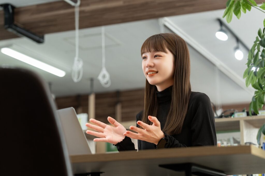 黒い服を着た女性がノートパソコンの前で話しながら身振り手振りを使って説明している様子。