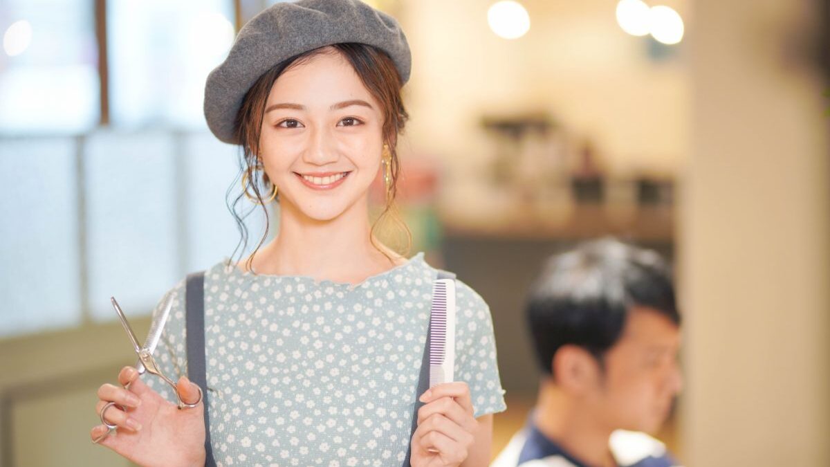 A young hairstylist smiling while holding scissors and a comb in a salon, representing a friendly and approachable junior hairstylist.