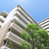 Exterior view of a mid-rise apartment building under a clear blue sky. Green trees in the foreground create a clean and safe impression.