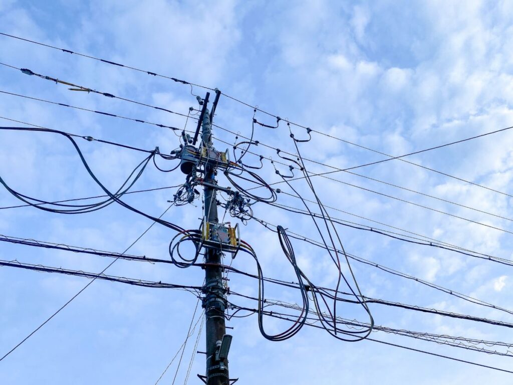青空の下に立つ電柱と複雑に張り巡らされた光回線ケーブル。強風によるケーブル損傷や通信障害のイメージ。