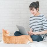 A woman sitting on a bed, working on a laptop while gently gazing at an orange cat lying at her feet.
